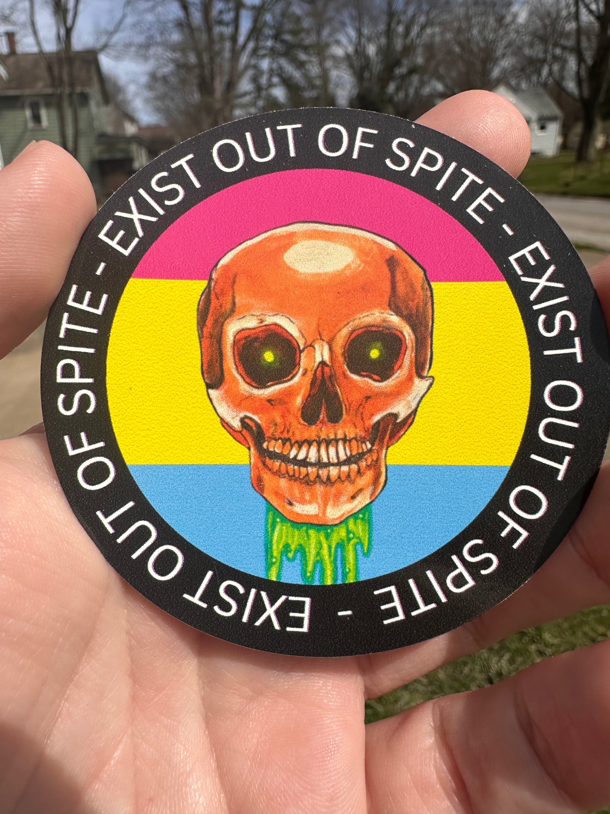 Round sticker with a skull over a pansexual pride flag encircled by the text exist out of spite