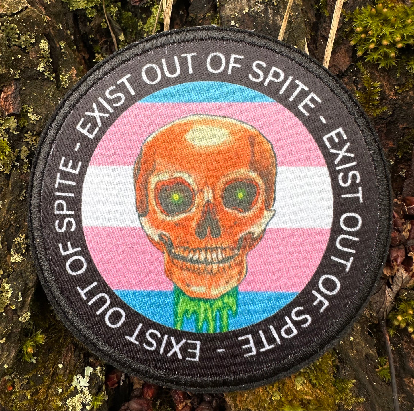 Patch with skull with transgender flag background circled with ‘exist out of spite’ text circling it