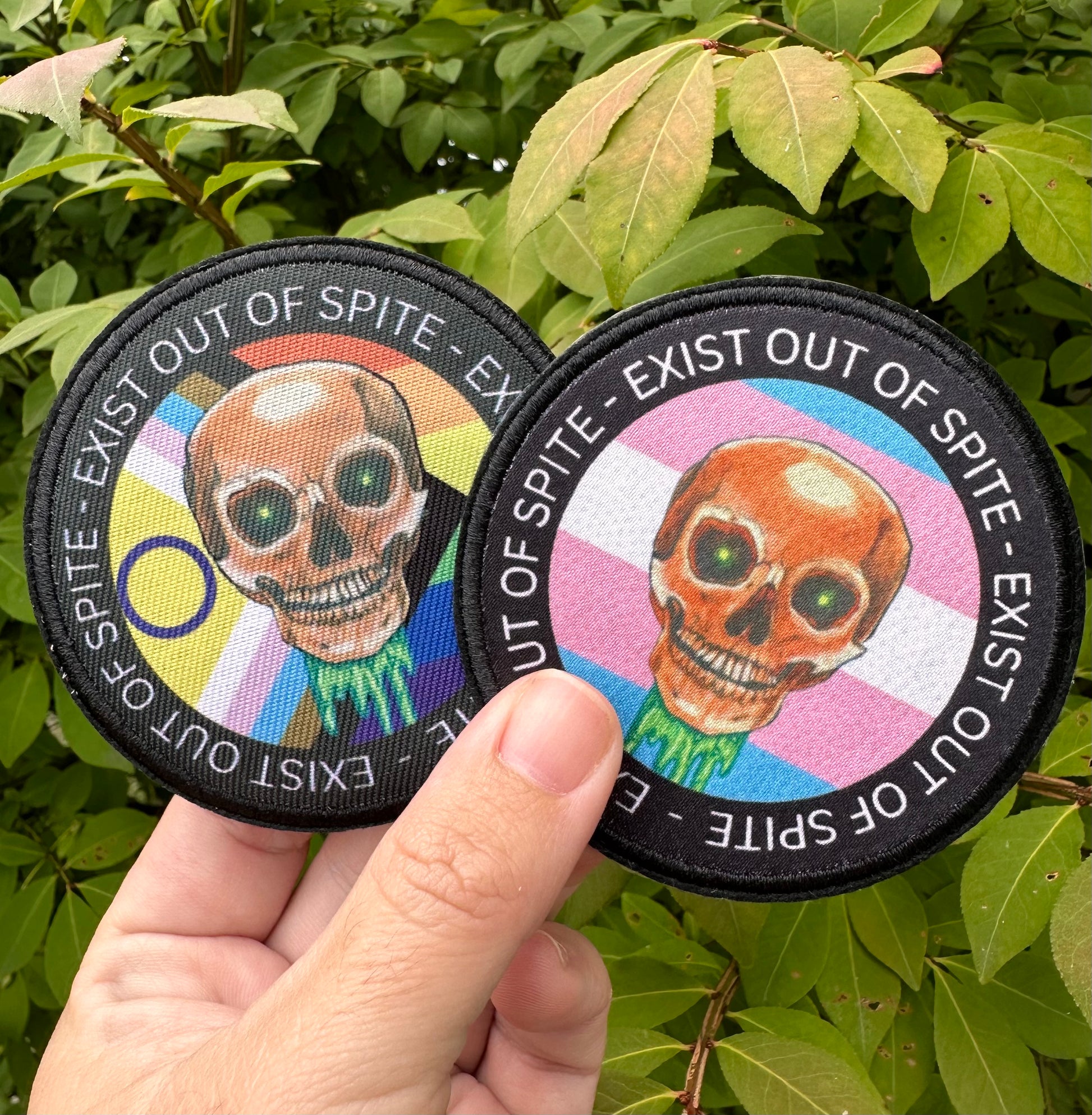 Two patches with a skull design, one with a pride flag one with a transgender flag held against a green leafy background