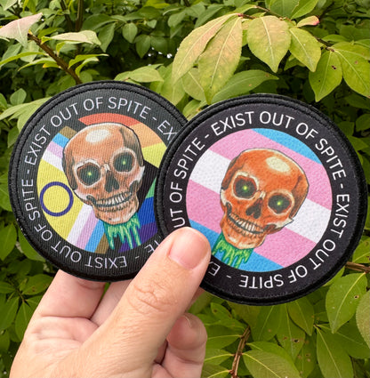 Two patches with a skull design, one with a pride flag one with a transgender flag held against a green leafy background