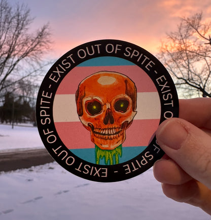 Round sticker with a skull over a transgender pride flag encircled by the text exist out of spite