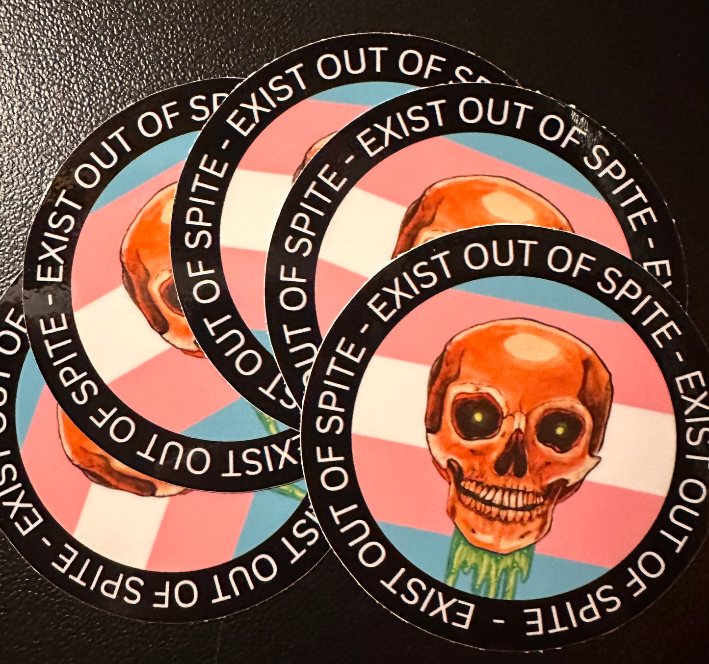 Round sticker 5 pack with a skull over a transgender pride flag encircled by the text exist out of spite