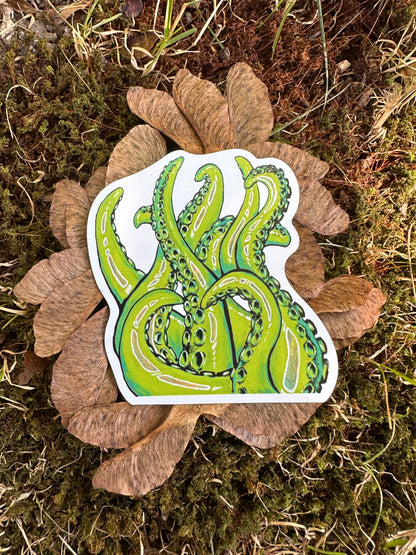 A magnet with a vibrant illustration of curling green tentacles on a white background, placed on a mossy surface with brown leaves scattered around.