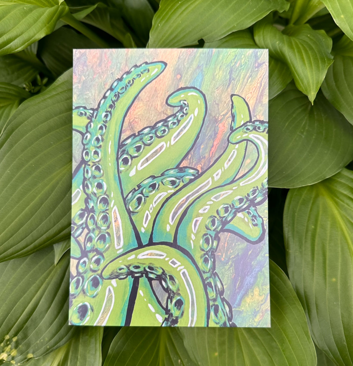 A colorful art print featuring an intricate illustration of tentacles on a glossy cardstock background, presented against a natural green leaf backdrop.