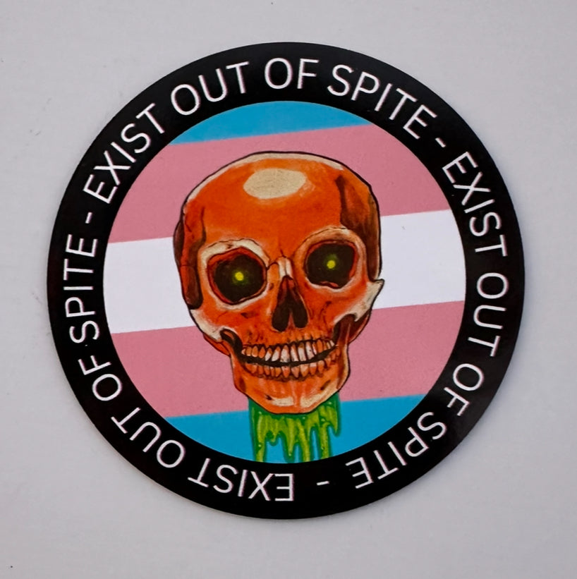 Round magnet with a skull over a transgender pride flag encircled by the text exist out of spite