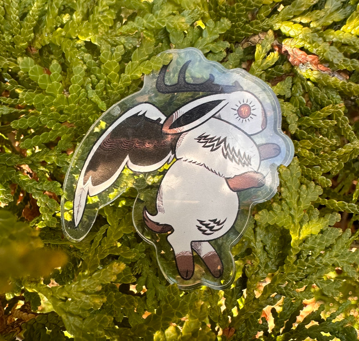 An acrylic pin depicting a whimsical creature with horns and wings, resembling a cross between a jackalope and a witch's familiar, set against a foliage background.