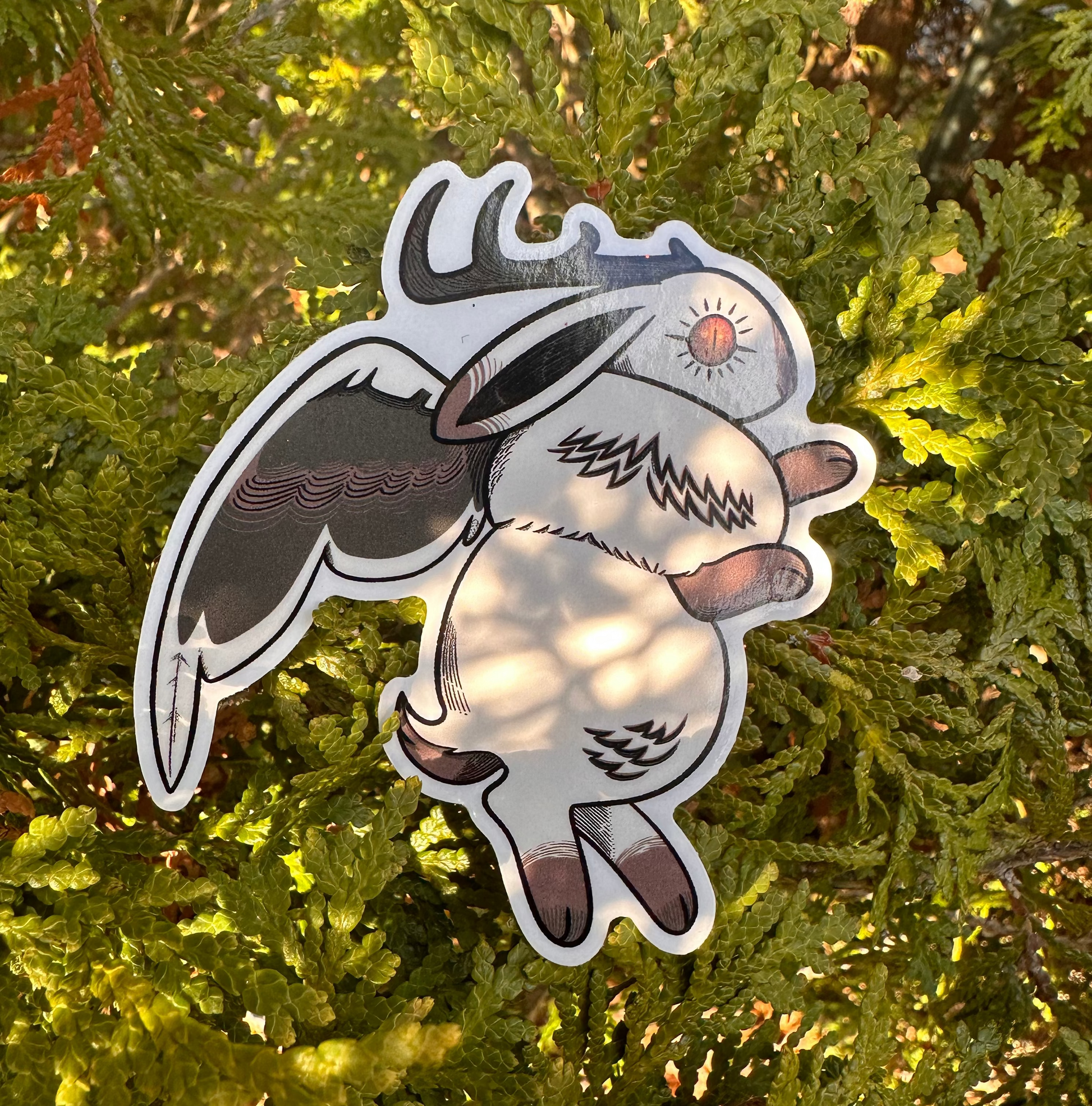 A vinyl sticker depicting a whimsical creature with horns and wings, placed against a foliage background.